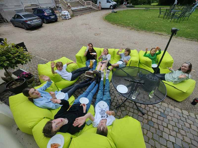 Viele Kinder mit fast leeren Pizzatellern sitzen, die Füße hochgelegt, draußen auf grünen Sitzsack-Sofas