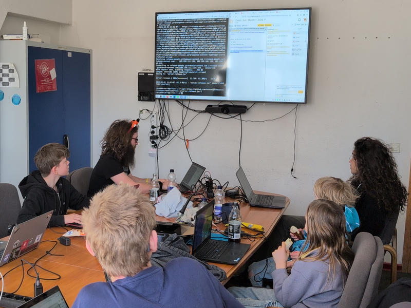 Kinder, Jugendliche und 
Erwachsene sitzen um einen Tisch neben ihren Laptops und schauen auf einen großen 
Bildschirm.