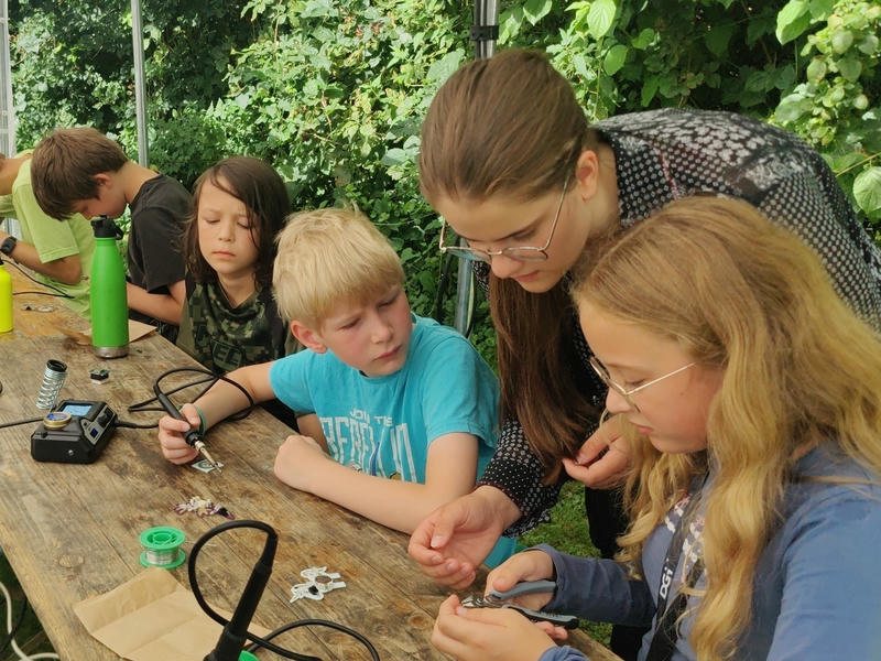 Mehrere Kinder sitzen an einem Tisch in einem Pavillon und lernen Löten