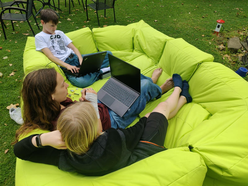 Drei Jugendliche sitzen mit Laptops auf einer Sitzsack-Sofa-Garnitur auf der Wiese, zwischen ihnen liegen einige Elektronikbauteile.