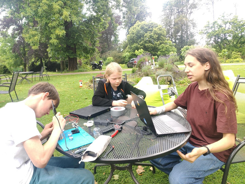 Drei Jugendliche sitzen draußen um einen Gartentisch. Einer lötet auf einer Lötunterlage, einer macht etwas an seinem Laptop.