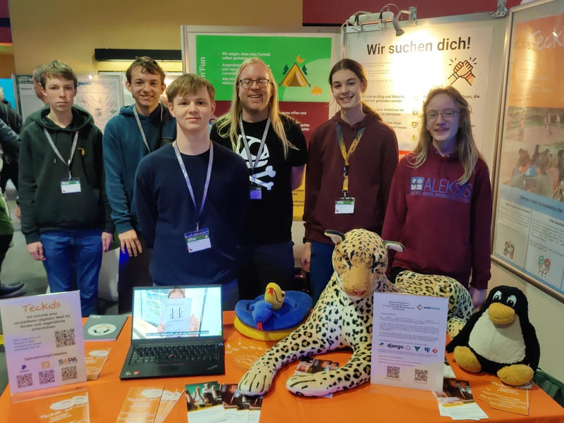 Florian, Robert, Lukas, Nik, Tuxilio und Pingu hinter einem orange gedeckten Infostand mit Plüsch-Leopard, Plüsch-Pinguin, einem Laptop und einigen Flyern